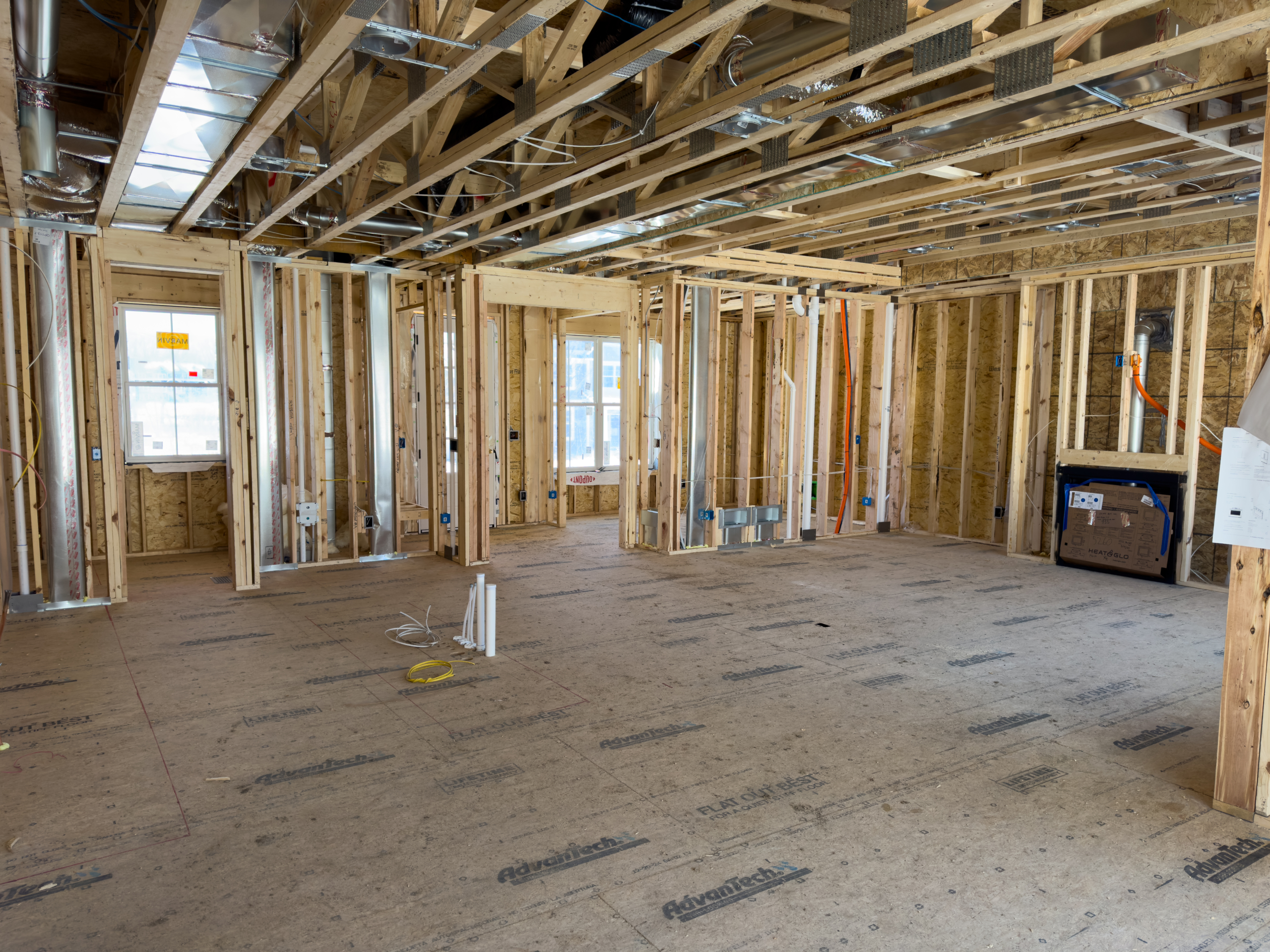 Open-concept gathering room of custom home at rough mechanicals phase showing exposed wood framing, HVAC ducts, electrical wiring, plumbing stubs, and AdvanTech subfloor